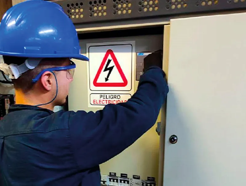 Técnico con casco y anteojos de seguridad abriendo un tablero eléctrico con señal de peligro eléctrico